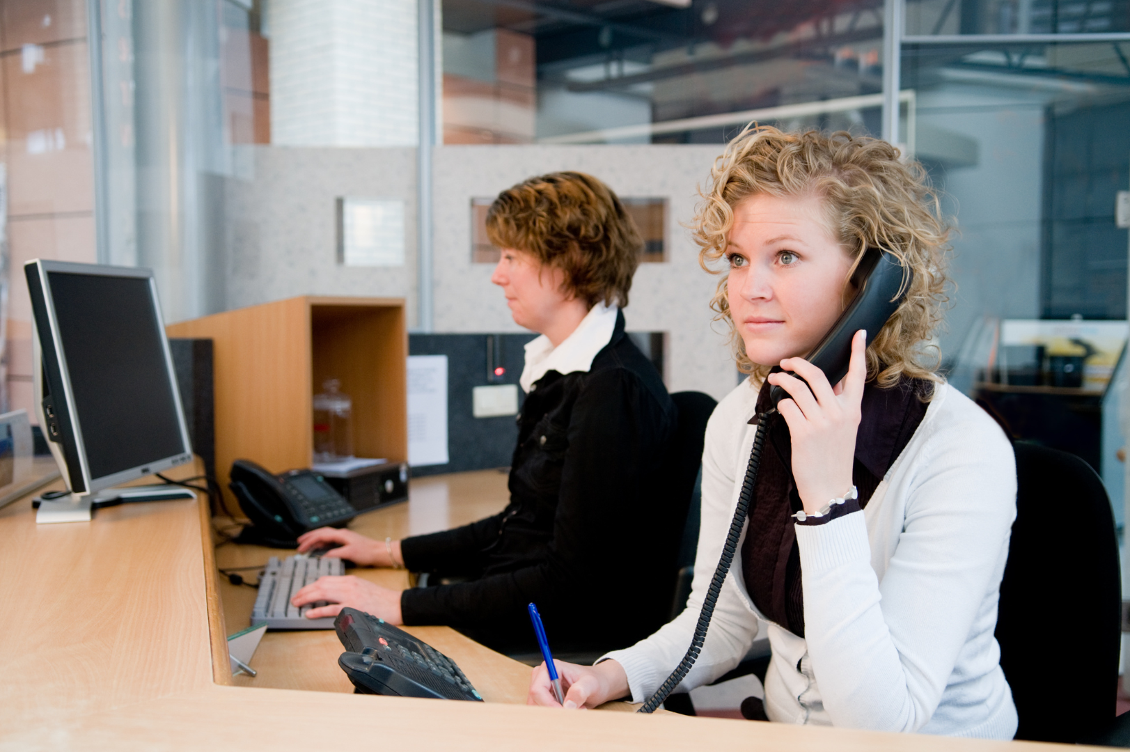 Woman at desk answering phone with a pen in her hand. Another woman behind her at the same desk working on the computer.